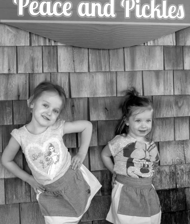 Two little girls in pickle-shop aprons posing under the Peace and Pickles sign